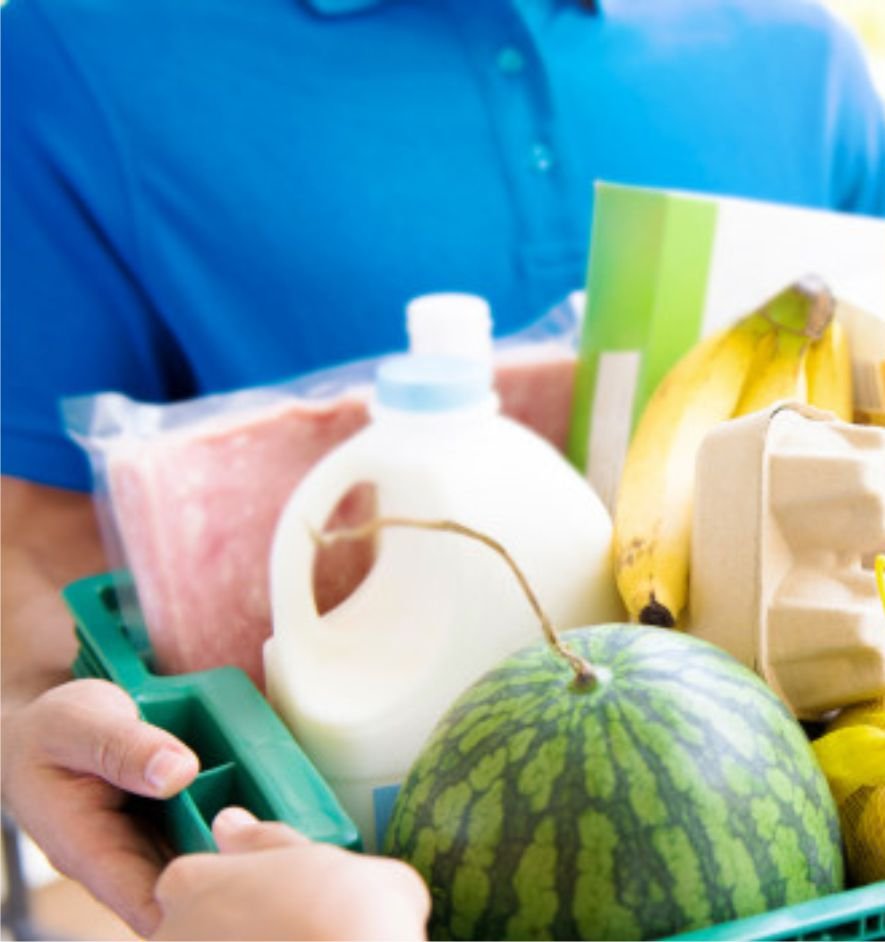 Caixa plástica verde segurada por uma pessoa de camisa polo azul, com carne embalada, um galão branco, uma melancia, bananas e uma caixa de ovos, e as mãos de outra pessoa pegando a caixa