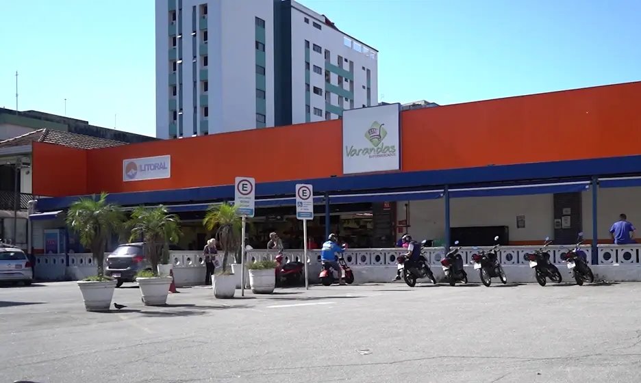 Loja do Varandas com fachada laranja, o letreiro da marca e uma placa da Rede Litoral, palmeiras em vasos, carros e motos estacionados e um prédio alto ao fundo